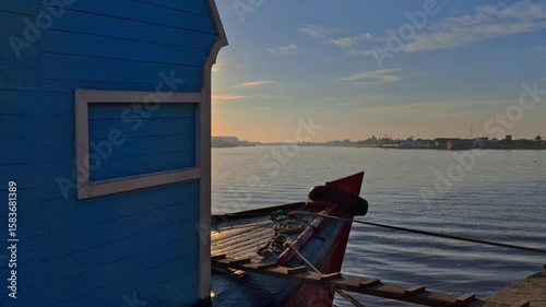 A traditional wooden boat with a blue cabin is moored on the river with the sun just rising above the horizon
