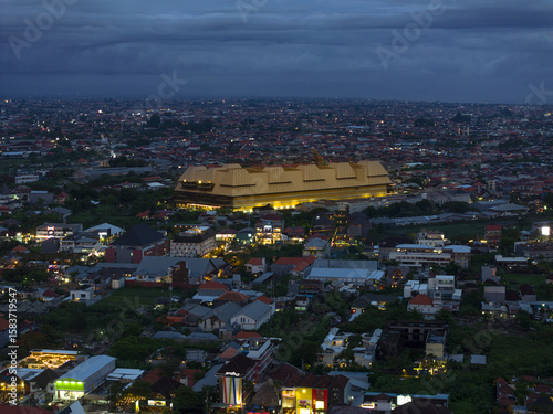 Aerial view of a sprawling urban landscape with a prominent, large, brightly lit building at dusk, its form stark against the darkening sky, Badung, Bali, Indonesia.