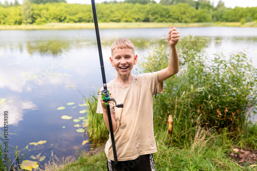 Happy teen boy holding fishing rod with caught fish on lake shore in summer