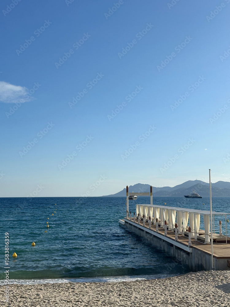 Fototapeta premium Serene beach scene with a wooden pier extending into calm blue waters, surrounded by gentle waves and distant mountains under a clear sky, evoking tranquility and relaxation