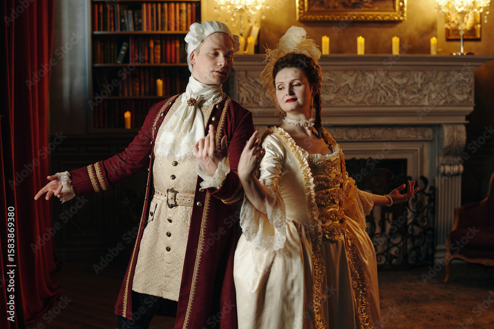 Obraz premium Caucasian young adult man and Caucasian young adult woman wearing Baroque period costumes performing elegant dance in ornate Renaissance style interior with bookshelves and fireplace