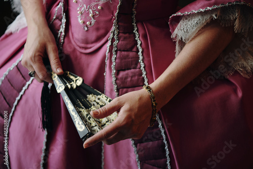 Caucasian middle aged woman holding ornate hand fan while wearing elaborate Baroque style dress with lace trim, hands positioned delicately in front of detailed historical costume