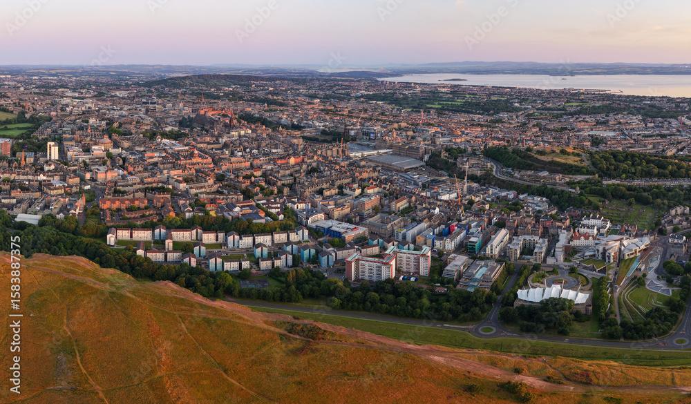 Fototapeta premium Panoramic Aerial image of the beautiful Edinburgh Cityscape featuring Holyrood Park touched by the first light of the morning Sunrise. 