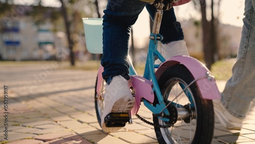 Canvas Print little child turns wheels on a two-wheeled bicycle, mother helps kid learn to control a bicycle, baby steers keeping balance, wheel and leg close-up, walk in the park mom and baby, road travel game