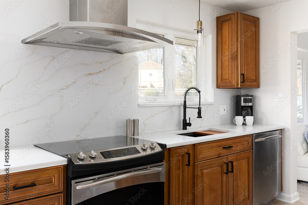Fototapeta premium A kitchen detail with a stainless steel stove and range hood, a white calacatta marble slab back splash and white countertop, wood cabinets, and a gold and black light hanging above the black faucet.