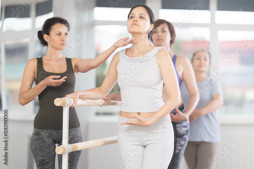 Positive focused young Asian woman refining ballet technique at barre with help of female instructor during beginner group lesson in choreography studio