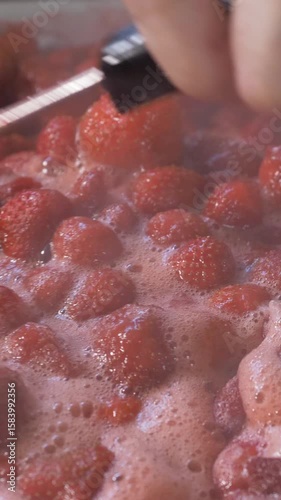 Boiling strawberries in a large pot release steam, with bubbles forming as they cook. A spoon is used to stir the mixture to keep it from sticking and to blend the ingredients