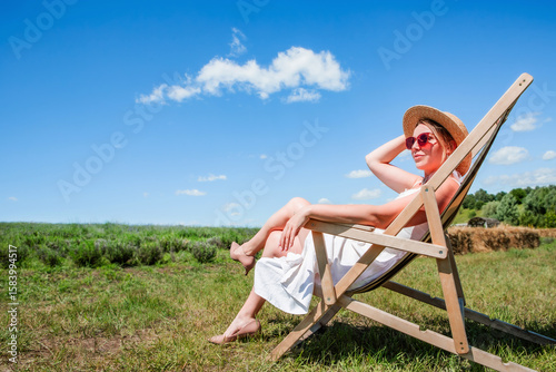 Young beautiful sexy woman in straw hat and sunglasses sitting on a sun lounger on a meadow on a sunny day against the sky. Rest. Vacation. Tourism.