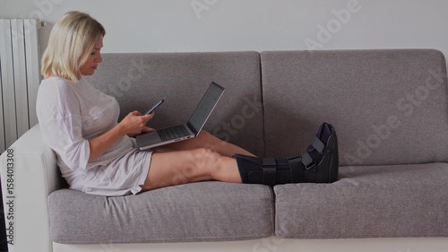 Woman with fractured leg sitting on the sofa with orthopedic medical Walker boot, splint foot, speaking on smartphones and working on the laptop. Remote jobs during the recovery from the trauma