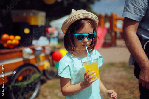 Little Girl Drinks Lemonade at a Kids Festival . Happy child enjoying summer vacation wearing fashionable outfit 
