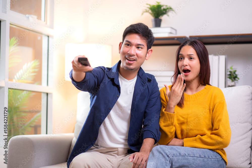 Obraz premium Young asian couple sitting on sofa in living room at home, reacting with surprise while watching television, expression emotional response during a dramatic scene, leisure together in cozy.