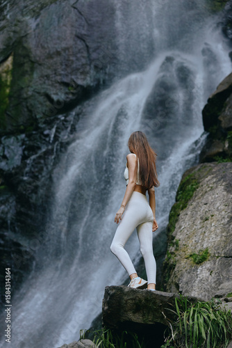 Woman in white activewear stands confidently on rock by a cascading waterfall surrounded by lush greenery in a serene natural setting during daytime.