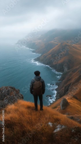 A person standing on a hilltop overlooking the ocean, with a backpack, wearing outdoor clothing. Dramatic clouds and vistas suggest an adventurous journey.