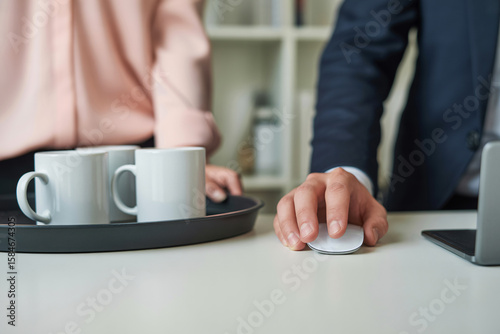 Close up of a woman’s hand in pastel pink blouse holding a tray with white coffee mugs next to a man’s hand on a large wireless mouse. Gender roles in the office