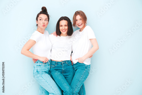 Three young beautiful smiling hipster female in trendy same summer white t-shirt and jeans clothes. Sexy carefree women posing near light blue wall in studio. Cheerful and positive models having fun