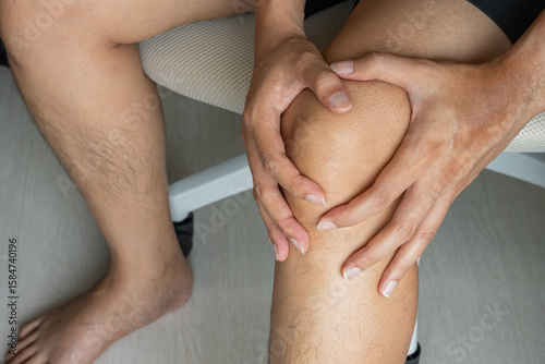 Obraz na plátně An Asian man sits on a cushioned chair, gripping his knee in apparent pain
