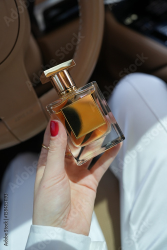In the car, a girl with a red manicure is holding a luxury perfume in a golden bottle. Hands are adorned with stylish accessories
