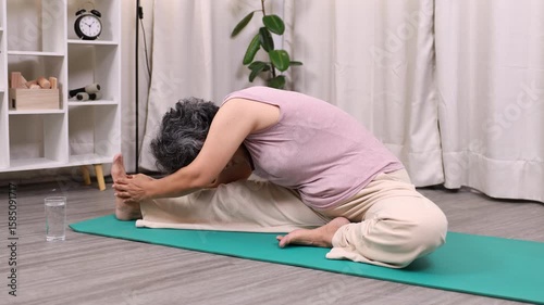 Senior Asian woman practicing a seated yoga stretch at home, bending forward with hands on her foot, embracing flexibility and mindfulness on a calm morning in a cozy home interior, yoga, meditation.