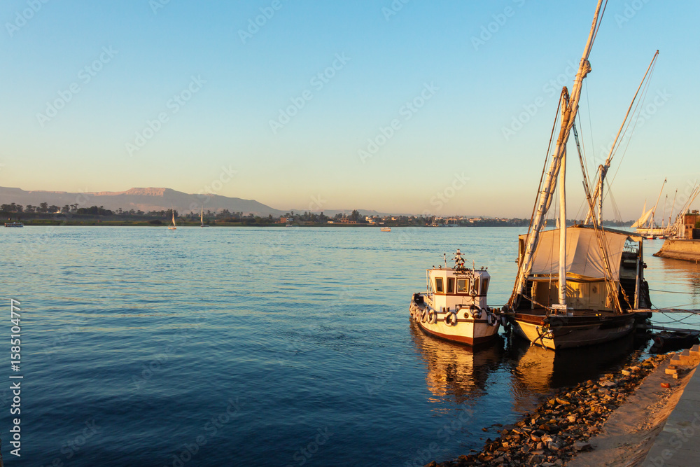 Fototapeta premium Traditional felucca boats moored on the Nile River at golden hour in Luxor, Egypt with mountain view