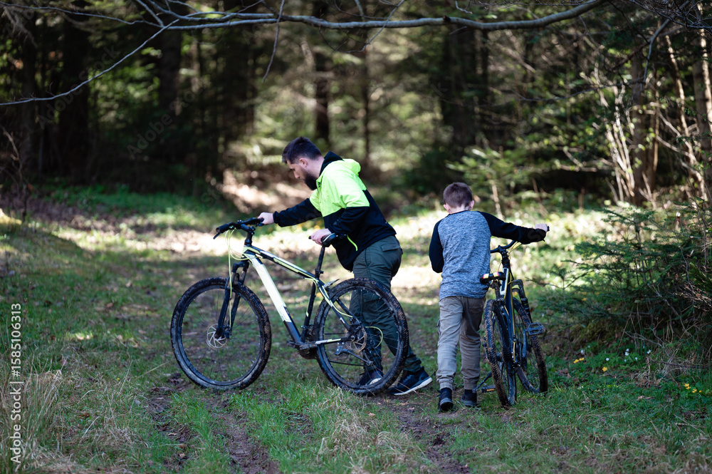 Fototapeta premium Father and son pushing muddy bikes on a forest trail. A sunny day of outdoor adventure.