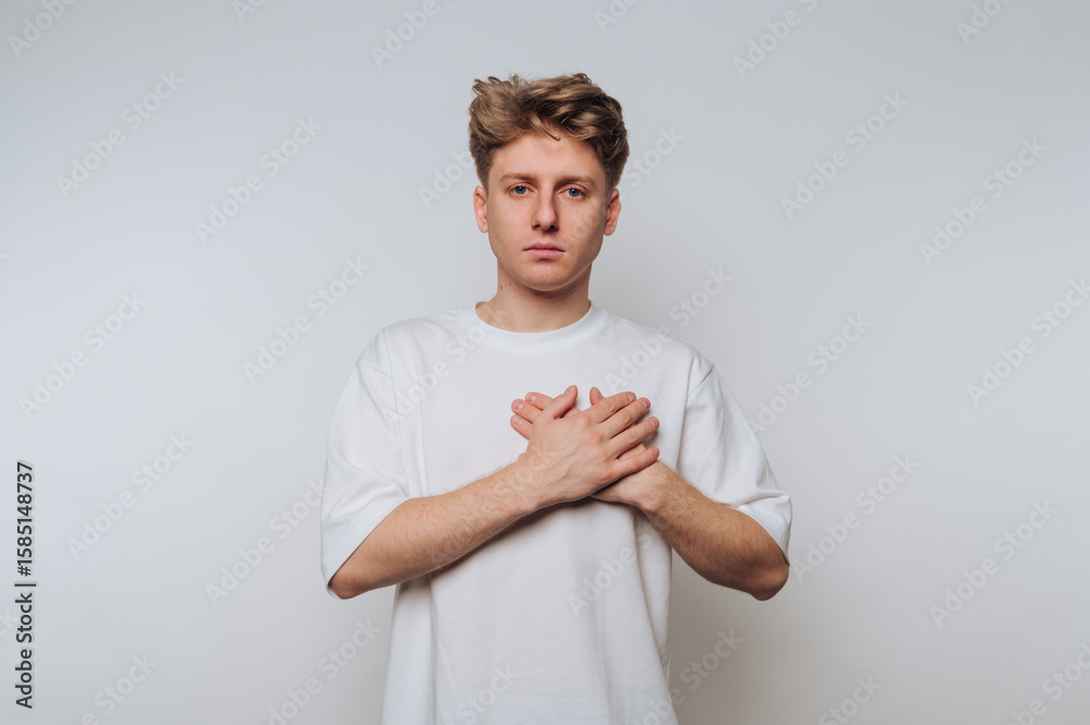 © bodnarphoto - Young man with neutral expression holds hands over chest against plain backdrop, conveying emotion and introspection