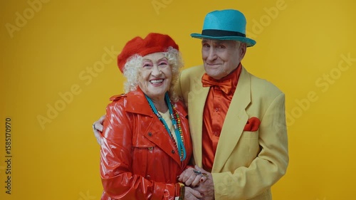 Senior couple radiating joy, sporting vibrant, colorful attire and sharing an affectionate moment while holding hands against striking yellow studio backdrop