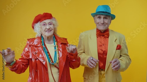 Senior couple dancing energetically in vibrant, colorful attire, radiating joy and vitality against bright yellow background while enjoying playful, lively moment together