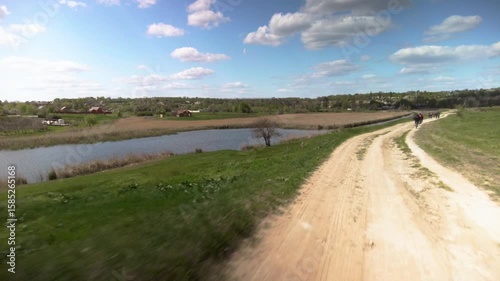 Group of active cyclist riding on meadow road by a river on summer day. Cyclists ride on dirt road along river in rural landscape. Adventure, nature and outdoor travel.
