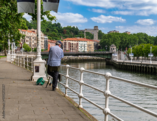 An unrecognizable man seen from behind fishing quietly on the riverside, with the City Hall bridge crossing the Nervión River visible in the background.