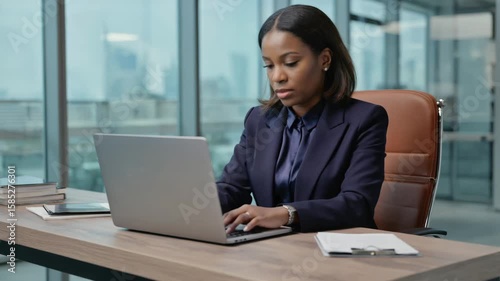 Professional black woman in suit using computer at desk in office with city view out window.