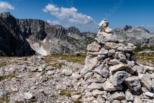 Stone cairn on a rocky alpine plateau with dramatic mountain peaks and patches of snow. Clear blue sky. Ideal for themes like hiking, trekking, adventure, navigation, or wilderness exploration.