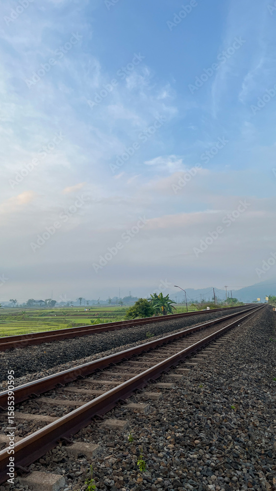 Fototapeta premium A railway track running through vast rice fields with misty morning sky and tropical greenery in a rural countryside setting.