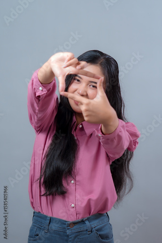 Indonesian woman in pink shirt making a creative hand frame gesture while smiling confidently. Studio portrait with casual style, long black hair, and playful, imaginative pose.
