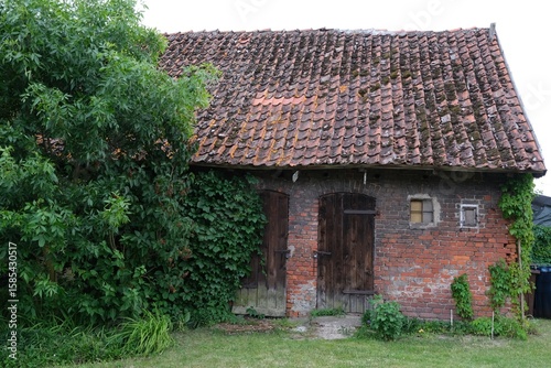 Old brick farm building with red roof tiles and wooden doors in countryside in Polish village, Kociewie. Green trees around.