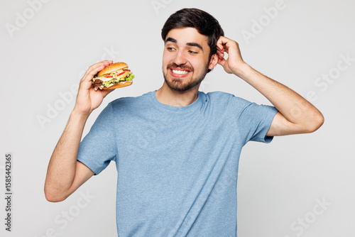 Young tempted sad minded confused man wears blue t-shirt casual clothes hold eat burger scratch head isolated on plain solid white background. Proper nutrition healthy food unhealthy choice concept.