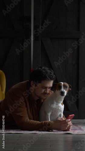 Vertical video of man with mobile phone and Jack Russell Terrier sitting on home floor, scrolling social media feed