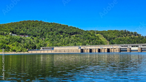 Modern dam and hydroelectric power station surrounded by lush green forest on a clear summer day. The facility is reflected in the calm surface of the reservoir under a bright blue sky. Sustainable en