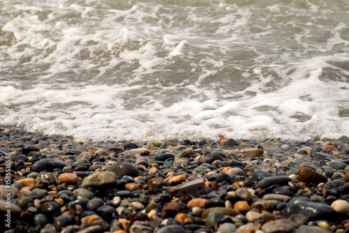 Foamy strong sea waves crashing on pebble beach on Black Sea coast at storm. Waves splashing on stones shore in stormy weather. Stormy water surface. Mystical seascape with waves broken on coastline.
