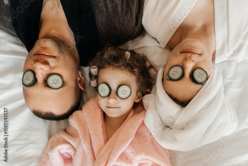 A family enjoys a spa day at home, with cucumber slices on their eyes, relaxing together on a white bed.