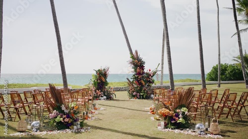 Tropical beach wedding setup with wooden chairs, palm trees, and vibrant floral arrangements by the ocean, ideal for destination or outdoor ceremonies. 
