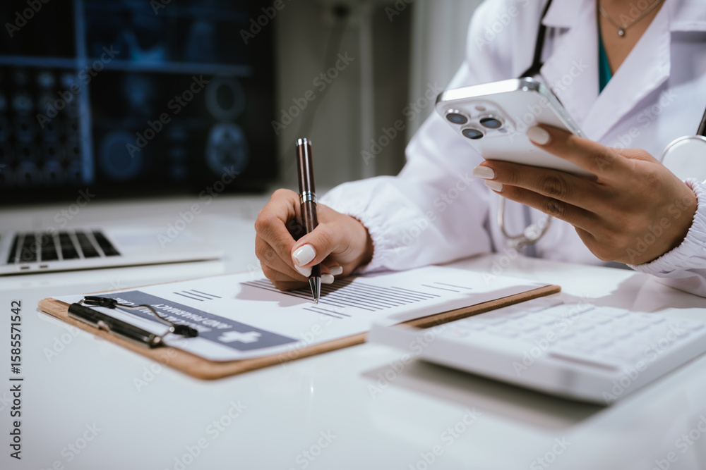Fototapeta premium Female doctor coordinating and writing on clipboard while holding smartphone, surrounded by modern devices such as laptop, calculator and diagnostic screen in medical facility.