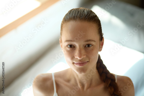 Free Download Skin-realism. Young woman smiles while sitting indoors with braided hair illuminated by soft natural light, showing natural pores, skin texture, and subtle shine. Concept of natural beauty, no makeup - GFXLoad