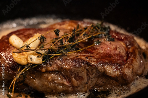 closeup of a rib-eye steak with garlic and thyme, cooking in butter on a cast iron skillet