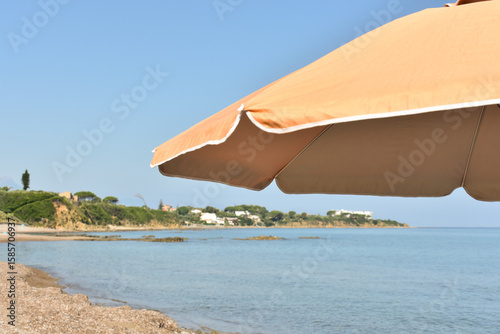 Fototapeta Naklejka Na Ścianę i Meble -  Parasol in the sand of the beach, summer and holidays. 
Summer beach in Italy and orange umbrella offering shade under the bright blue sky. Holidays in Sicily. Beach near Cefalu. Travel to Italy. 