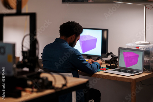 An African American man works on a 3D design project in his workshop. He manipulates a component, with two computer screens displaying a purple cube model.