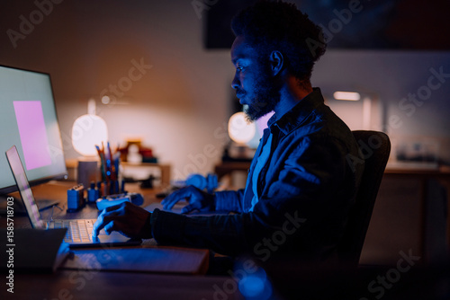 An African American man works intently on his laptop and large monitor in a dimly lit home office. He is focused on his task, illuminated by screen light.