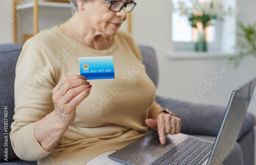Old senior mature bank client woman making online payment holding plastic card, credit card, paying for purchase on Internet, using application, shopping at home, elder financial study banking service