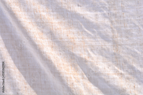 A close-up view of a light-colored, slightly textured fabric with subtle wrinkles and hints of natural fibers, illuminated by sunlight