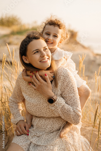 A loving mother and her happy daughter share a tender moment in sun-kissed tall grasses, both smiling.