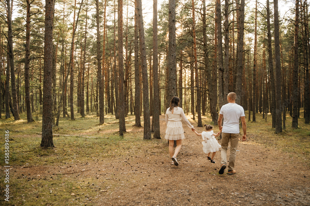 Fototapeta premium A happy family of mother, father, and daughter walks hand-in-hand away from the camera on a sun-dappled forest path.